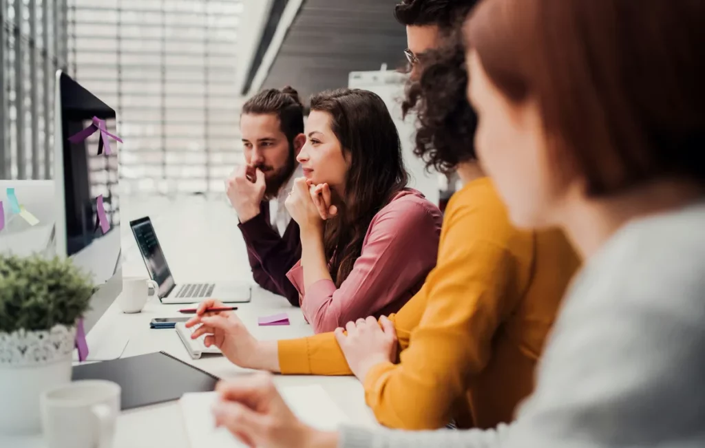 Trois collègues de travail échangent et sourient autour d’un ordinateur portable dans un bureau moderne, reflétant la collaboration et la convivialité des partenariats professionnels.